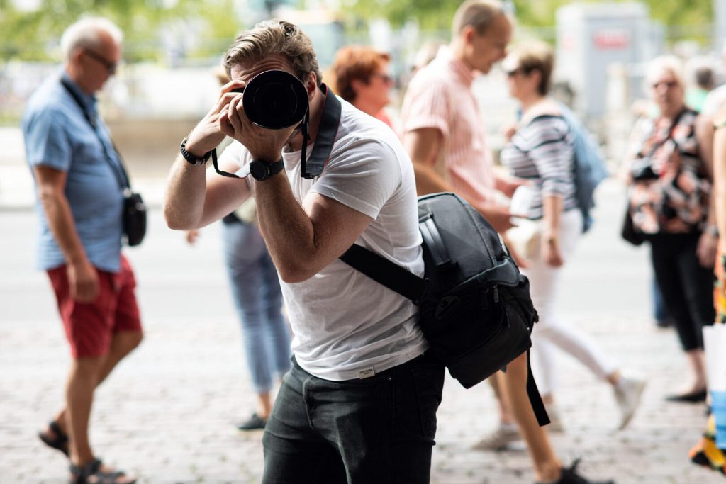 En person med et kamera tager et billede på en travl gade og lægger dygtigt strategier for at finde det perfekte billede. Andre mennesker går forbi, nogle med tasker eller kameraer, der hver især tilføjer deres unikke touch til den urbane baggrund.