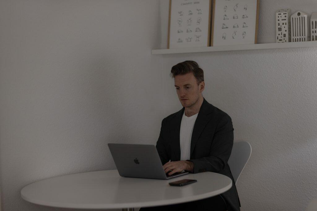 En mand i mørkt jakkesæt sidder ved et rundt hvidt bord og bruger en bærbar computer til at forfine sin brandingstrategi med en smartphone i nærheden. Bag ham viser en hylde indrammet minimalistisk kunst og små hvide modeller, der forstærker det slanke billede af hans arbejdsområde.