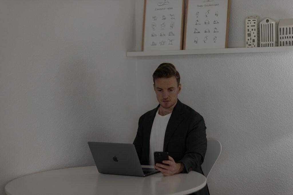 En mand sidder ved et rundt bord, strategisk ved hjælp af en bærbar computer og smartphone. Indrammede kunst- og modelbygninger afspejler subtilt hans skarpe øje for branding, hvilket forstærker billedets dybde i baggrunden.