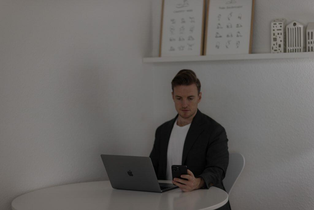 En person i en mørk jakke sidder ved et rundt hvidt bord med en bærbar computer og tjekker deres telefon. Rummet, der inkarnerer en brandingstrategi af minimalisme, byder på indrammet kunst og små bygningsmodeller på en hylde.