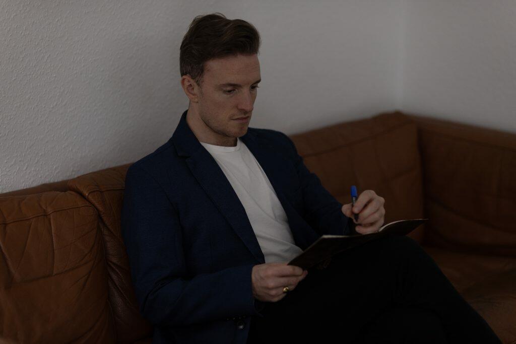 En person i en blå blazer og hvid skjorte sidder på en brun sofa, holder en kuglepen og en notesbog, fordybet i at udvikle brandingstrategier.