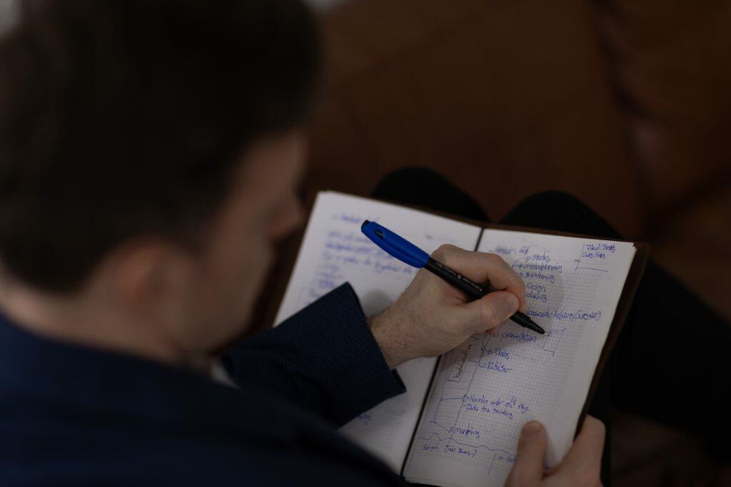 Siddende på en brun sofa skriver en person strategiske noter og diagrammer i en notesbog med en blå pen og omhyggeligt udforme ideer, der vil forme deres personlige brand.