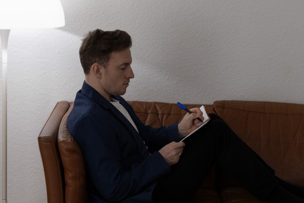 En mand sidder på en brun sofa og planlægger sit næste træk, mens han skriver i en notesbog med en blå pen. Klædt i en marineblå blazer og hvid skjorte udstråler han professionalisme ved det bløde skær fra en hvid gulvlampe.