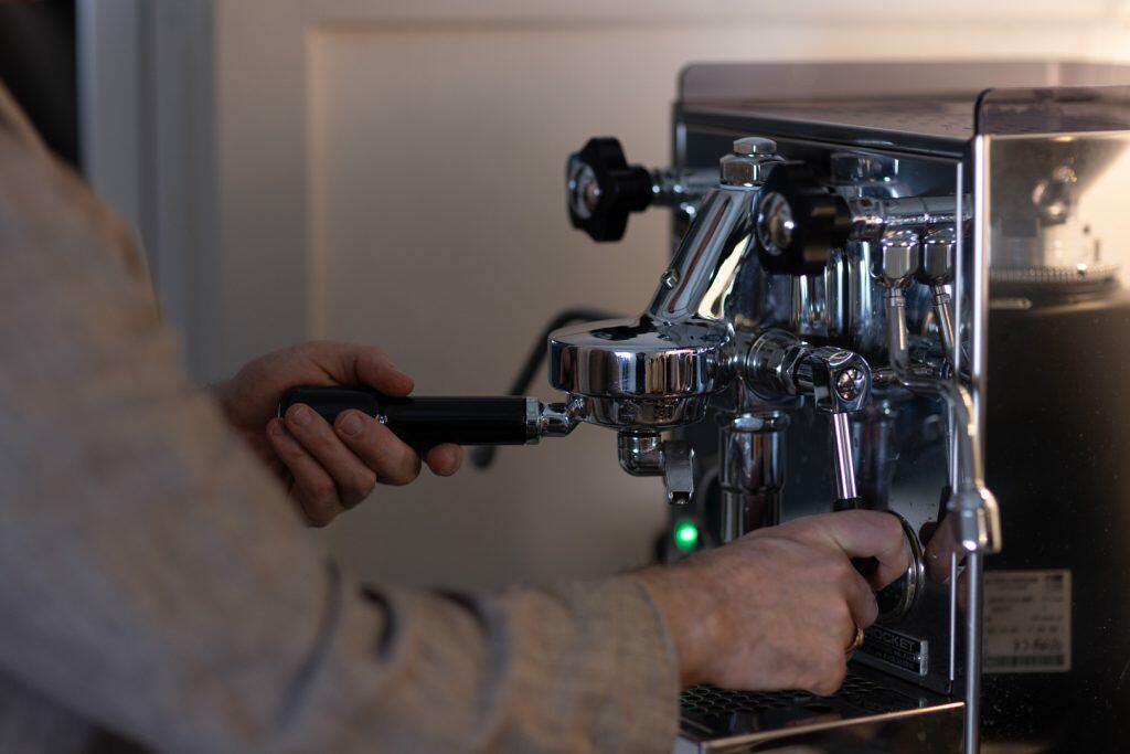 En person betjener dygtigt en espressomaskine, justerer portafilterhåndtaget, mens han fremhæver maskinens slanke metalelementer for at forbedre brandets image.