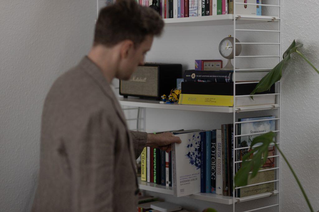 En person i en brun jakke vælger en bog fra en hvid bogreol fyldt med forskellige bøger og dekorative genstande, og skaber deres personlige billede med hvert tankevækkende valg.