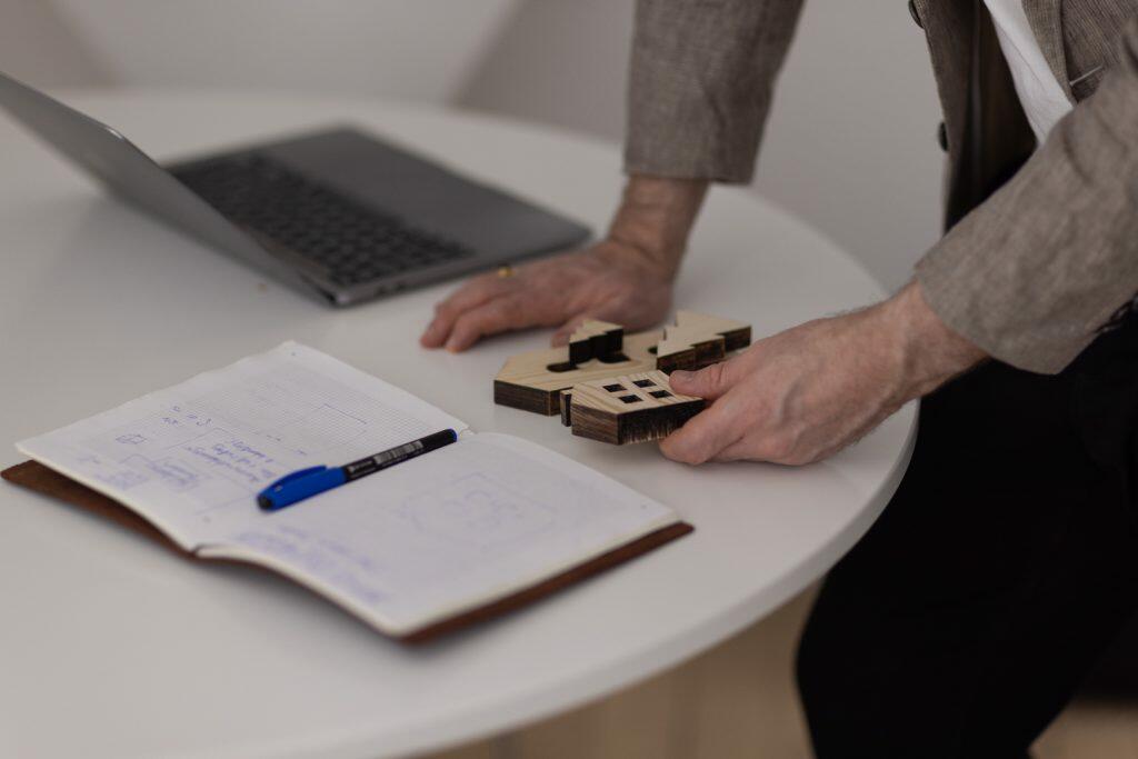 En person samler strategisk en puslespilsbrik i træ på et rundt hvidt bord ved siden af en åben notesbog med skitser og en kuglepen og et billede på deres bærbare computer.