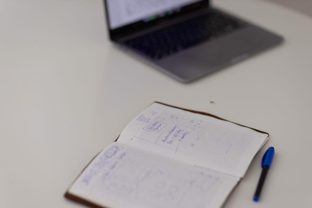 En åben notesbog med håndskrevne strategier og en blå pen hviler på bordet, mens en sløret bærbar computer i baggrunden subtilt forbedrer billedet af fokuseret produktivitet.