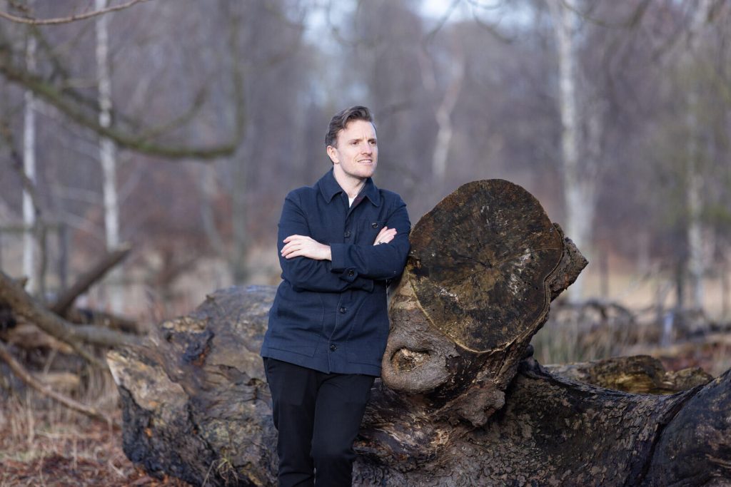 Klædt i et mørkeblåt outfit læner en person sig strategisk op ad en stor træstub i skoven, med arme over kors og øjne, der overvåger scenen. Sparsomme træer og nedfaldne grene danner et billede, der harmonerer med deres stille styrke, en ideel ramme for personlig branding.