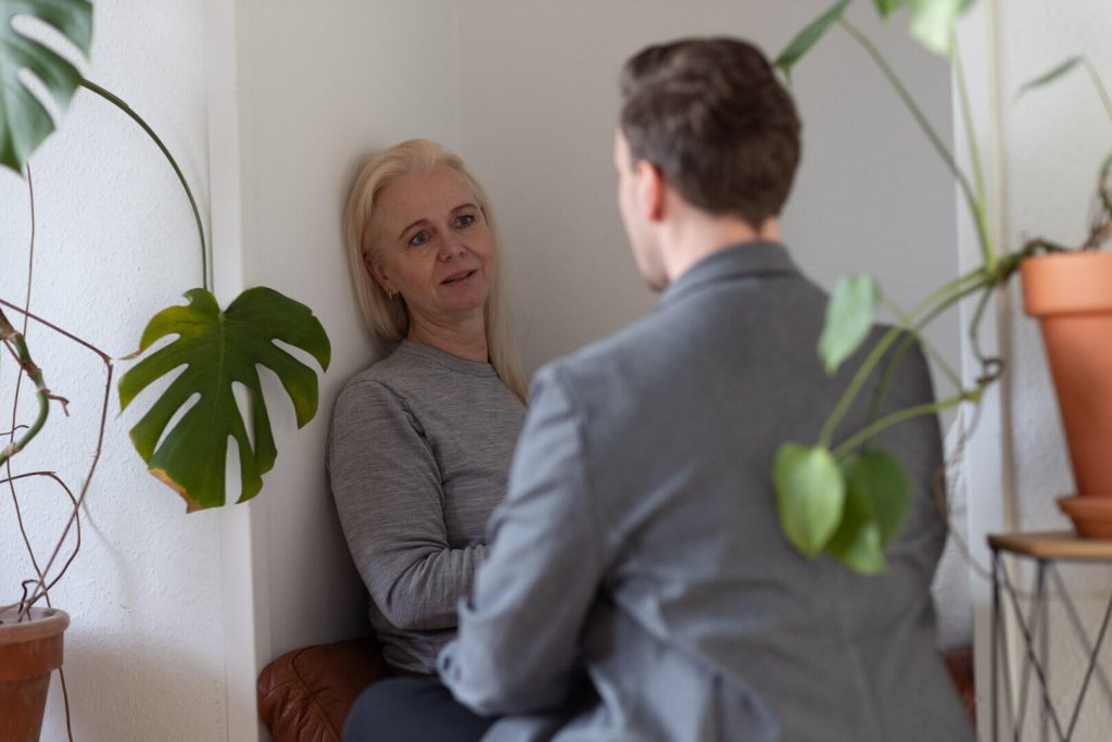 En kvinde og en mand sidder over for hinanden i et hjørne af et værelse, omgivet af potteplanter. Kvindens blik er strategisk, som om hun planlagde det næste skridt i deres branding-samtale.