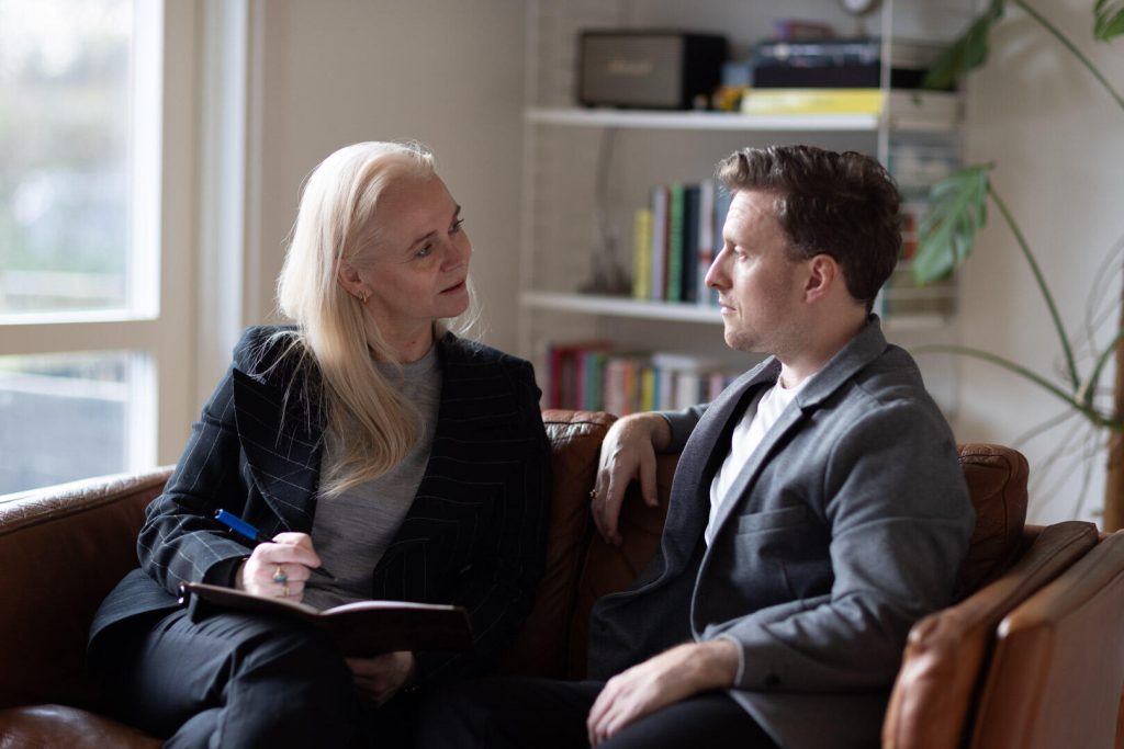 En kvinde med en notesblok og en mand sidder på en sofa, engageret i samtale om brandingstrategier. Hylder og et vindue er i baggrunden.