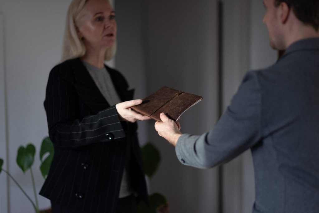 I et kontormiljø engagerer to personer sig i en strategisk udveksling af en brun mappe, den ene udvider den til den anden. En plante er synlig i baggrunden, hvilket forstærker billedets branding med et strejf af naturlig elegance.