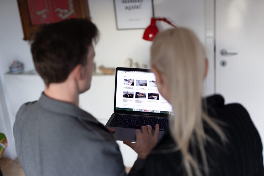 To personer lægger strategi over en bærbar computers skærm og viser adskillige miniaturebilleder i et rum med en rød lampe og en lukket dør.