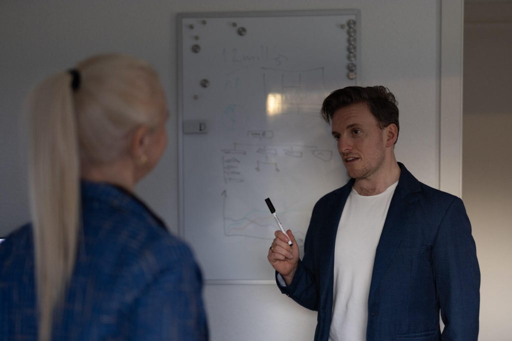 En mand, der holder en markør, diskuterer brandingstrategier med en kvinde foran en tavle fyldt med diagrammer og noter. Begge er klædt i forretningsdragt og projicerer et professionelt image.