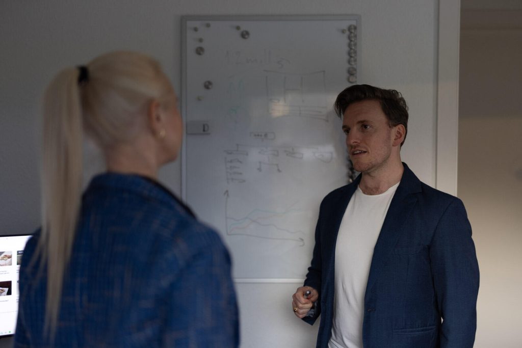 To personer i et rum har en strategisk diskussion foran en tavle fyldt med diagrammer. Personen til højre, der holder en markør, skitserer ideer for at forbedre deres brandimage.